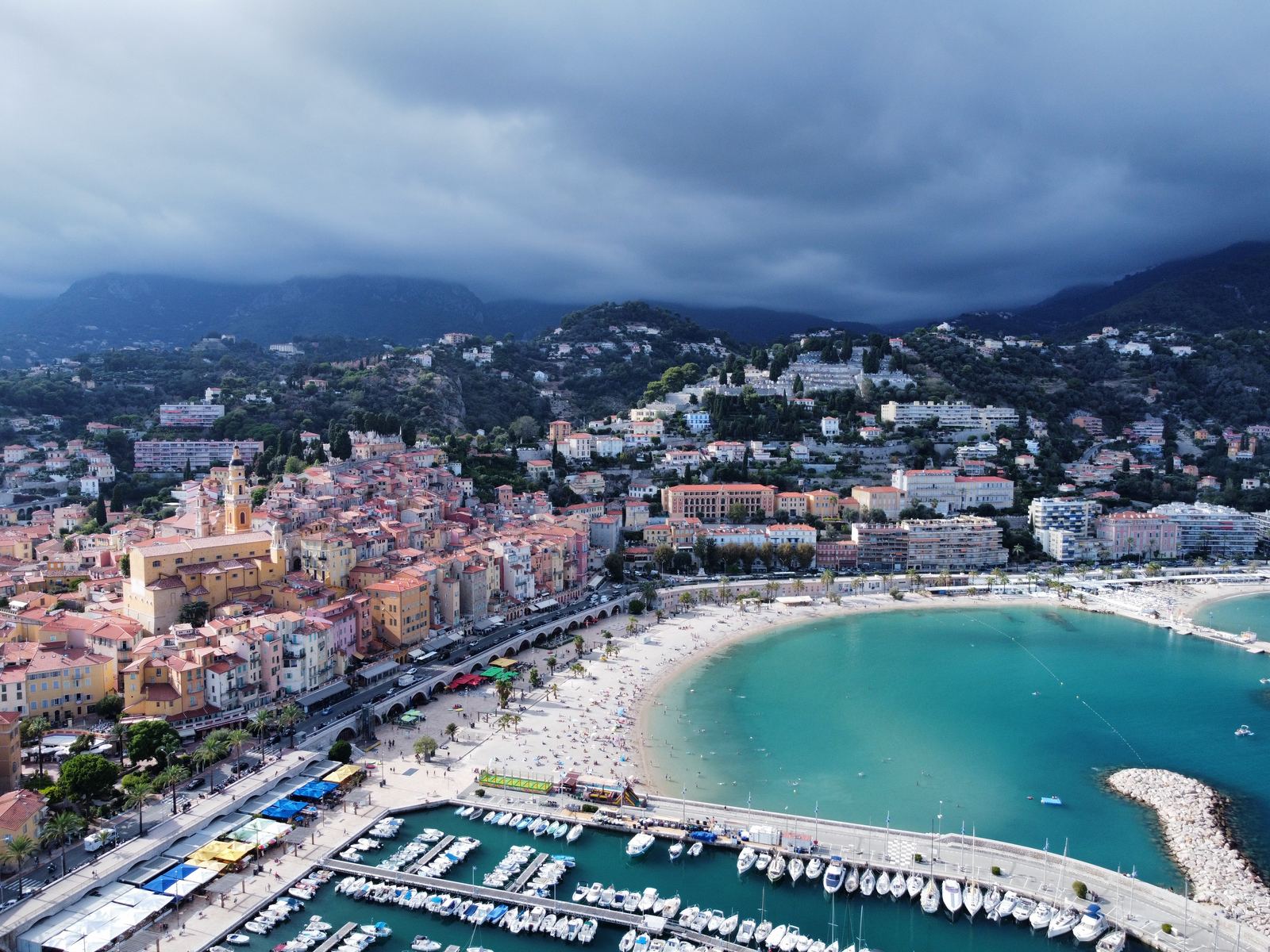 Menton from above — colourful old town, turquoise bay, the beach, the harbour, mountains with storm clouds behind